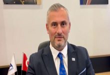 Orta yaşlı bir erkek, resmi bir ortamda masa başında oturuyor. Lacivert takım elbise, beyaz gömlek ve mavi kravat takmış. Saçları kısa ve griye dönmüş, yüzünde hafif sakal var. Arkasında Türk bayrağı ve bir kurum bayrağı görünüyor; genel atmosfer bir ofis ya da resmi kurum ortamını andırıyor.