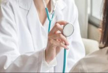 Beyaz önlüklü bir doktor, elinde stetoskopla bir hastayı muayene etmeye hazırlanıyor. Kadrajda doktorun üst kısmı ve hastanın omuz bölgesi görülüyor. Sahne, rutin bir sağlık kontrolü anını yansıtıyor.