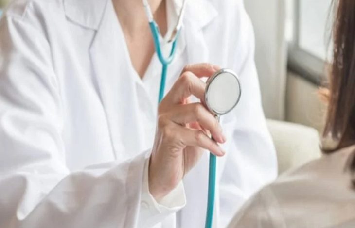 Beyaz önlüklü bir doktor, elinde stetoskopla bir hastayı muayene etmeye hazırlanıyor. Kadrajda doktorun üst kısmı ve hastanın omuz bölgesi görülüyor. Sahne, rutin bir sağlık kontrolü anını yansıtıyor.