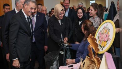 Resmi bir heyetin, el emeği ürünlerin sergilendiği bir masayı ve bu ürünleri yapan engelli bir bireyi ziyaret ettiği andır. Katılımcıların güler yüzle ürünleri incelediği, sosyal bir etkileşim ve sergi anı betimlenmektedir.