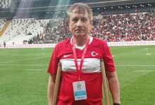 Bir stadyumda, koltuk değnekleriyle saha kenarında duran bir adam, üzerinde Türk bayrağı bulunan milli takım formasıyla poz veriyor. Boynundaki akreditasyon kartı ve arka plandaki tribünler, önemli bir spor organizasyonunun atmosferini yansıtıyor. Sahaya bazen ayakla değil, yürekle çıkılır.