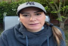 Açık havada, yeşilliklerin önünde oturan bir kadın yakın planda görünüyor. Gri şapka ve koyu renk kapüşonlu bir üst giymiş. Saçları arkadan toplanmış, kulaklarında kablosuz kulaklık var. Yüz ifadesi ciddi ve doğrudan kameraya bakıyor.