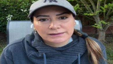 Açık havada, yeşilliklerin önünde oturan bir kadın yakın planda görünüyor. Gri şapka ve koyu renk kapüşonlu bir üst giymiş. Saçları arkadan toplanmış, kulaklarında kablosuz kulaklık var. Yüz ifadesi ciddi ve doğrudan kameraya bakıyor.