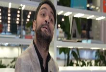 Modern bir iç mekânda, rafların ve bitkilerin önünde duran sakallı bir adam hafif yukarı bakarak konuşur gibi bir ifade sergiliyor. Üzerinde açık renk ceket ve koyu gömlek var; ortam aydınlık ve düzenli görünüyor.