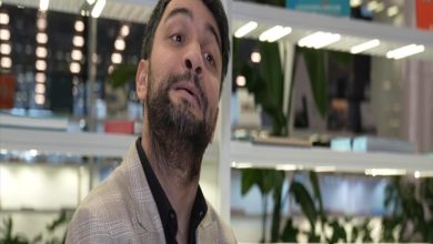 Modern bir iç mekânda, rafların ve bitkilerin önünde duran sakallı bir adam hafif yukarı bakarak konuşur gibi bir ifade sergiliyor. Üzerinde açık renk ceket ve koyu gömlek var; ortam aydınlık ve düzenli görünüyor.