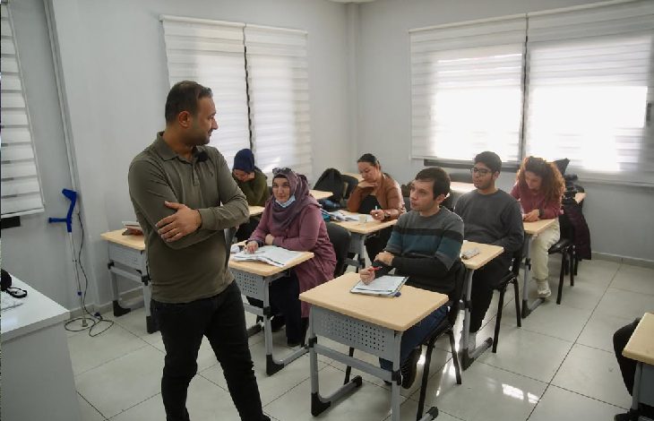 Bir sınıfta EKPSS hazırlık kursu alan öğrencileri ve başlarında duran bir eğitmeni gösteren fotoğraftır. Sınıftaki öğrenciler sıralarında ders çalışırken, eğitmen onlarla ilgilenmektedir. Odaklanmış ve eğitici bir atmosfer hakimdir.