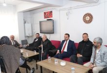 Sade bir oturma odasında, karşılıklı koltuklarda oturan bir grup insan sohbet ediyor. Takım elbiseli ve resmi görünümlü kişiler ile ev sahipleri bir arada. Masaların üzerinde çay bardakları var. Duvara asılı Türk bayrağı görseli ve saat dikkat çekiyor. Atmosfer, bir ziyaret ve samimi bir görüşme havası taşıyor.