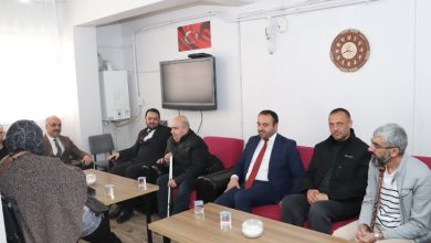 Sade bir oturma odasında, karşılıklı koltuklarda oturan bir grup insan sohbet ediyor. Takım elbiseli ve resmi görünümlü kişiler ile ev sahipleri bir arada. Masaların üzerinde çay bardakları var. Duvara asılı Türk bayrağı görseli ve saat dikkat çekiyor. Atmosfer, bir ziyaret ve samimi bir görüşme havası taşıyor.