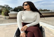 Görselde, dış mekânda tekerlekli sandalyesinde oturan bir kadın görülüyor. Beyaz, uzun kollu bir kazak ve bordo pantolon giymiş; güneş gözlüğü takıyor ve yana doğru bakarak sakin, kendinden emin bir duruş sergiliyor. Arka planda açık hava, ağaçlar ve mimari detaylar yer alıyor.