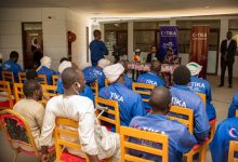 Bir grup insan, Türk İşbirliği ve Koordinasyon Ajansı (TİKA) logolu kıyafetlerle otururken, ön tarafta yapılan bir konuşmayı dikkatle dinledikleri resmi bir toplantı ortamında bulunuyor.