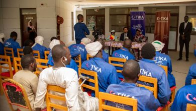 Bir grup insan, Türk İşbirliği ve Koordinasyon Ajansı (TİKA) logolu kıyafetlerle otururken, ön tarafta yapılan bir konuşmayı dikkatle dinledikleri resmi bir toplantı ortamında bulunuyor.