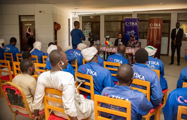 Bir grup insan, Türk İşbirliği ve Koordinasyon Ajansı (TİKA) logolu kıyafetlerle otururken, ön tarafta yapılan bir konuşmayı dikkatle dinledikleri resmi bir toplantı ortamında bulunuyor.