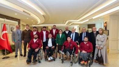 Şık bir salon ortamında, arka planda Türk bayrağının yer aldığı bir mekânda çekilmiş toplu bir fotoğraf. Önde tekerlekli sandalyede oturan sporcular (kırmızı eşofmanlı), arkada ise takım üyeleri ve resmi görünümlü kişiler yan yana poz veriyor. Genel atmosfer resmi ama samimi; bir başarı ya da ziyaret anı gibi duruyor.