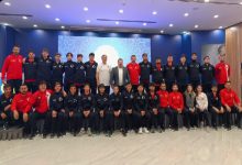 Kalabalık bir spor kafilesi, kapalı bir salonda toplu fotoğraf için poz vermiş. Sporcuların çoğu lacivert eşofman, bazıları kırmızı üstler giymiş. Ortada teknik ekip yer alıyor; arka planda büyük bir ekran bulunuyor.