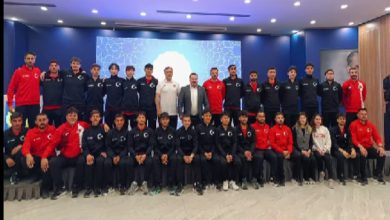 Kalabalık bir spor kafilesi, kapalı bir salonda toplu fotoğraf için poz vermiş. Sporcuların çoğu lacivert eşofman, bazıları kırmızı üstler giymiş. Ortada teknik ekip yer alıyor; arka planda büyük bir ekran bulunuyor.