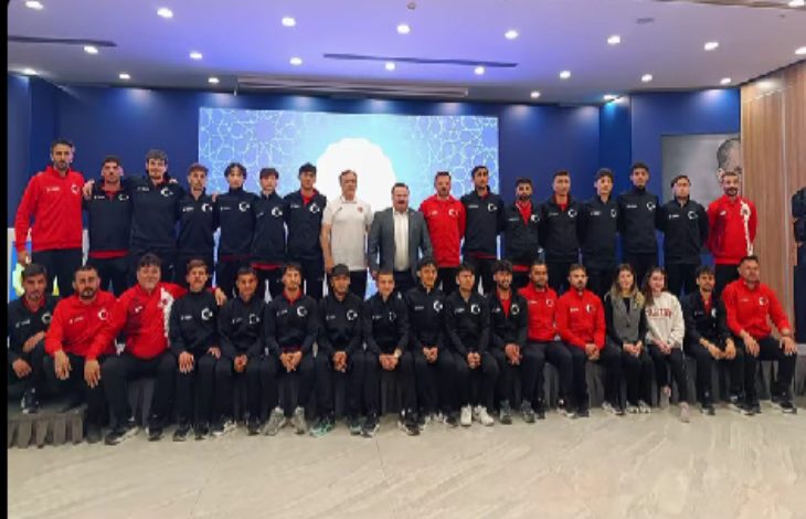 Kalabalık bir spor kafilesi, kapalı bir salonda toplu fotoğraf için poz vermiş. Sporcuların çoğu lacivert eşofman, bazıları kırmızı üstler giymiş. Ortada teknik ekip yer alıyor; arka planda büyük bir ekran bulunuyor.