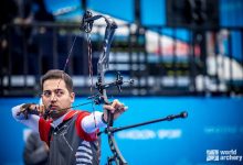 Milli para okçu Yiğit Caner Aydın'ın müsabaka esnasında hedefe odaklandığı ve yayını gerdiği anı gösteren aksiyon dolu bir kare. Aydın, profesyonel ekipmanıyla atış pozisyonunda görülürken, arka planda mavi renklerin hakim olduğu bir spor alanı atmosferi yer alıyor.