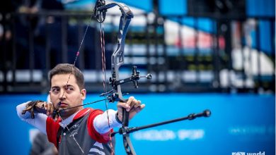 Milli para okçu Yiğit Caner Aydın'ın müsabaka esnasında hedefe odaklandığı ve yayını gerdiği anı gösteren aksiyon dolu bir kare. Aydın, profesyonel ekipmanıyla atış pozisyonunda görülürken, arka planda mavi renklerin hakim olduğu bir spor alanı atmosferi yer alıyor.