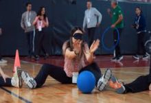 Bir spor salonunda, gözleri siyah bir bantla kapatılmış bir kadının yerde oturarak mavi bir topla paralimpik spor denemesi yaptığı anı gösteren bir fotoğraf. Arka planda etkinliği izleyen ve destek veren diğer katılımcılar ile spor ekipmanları görülüyor. Görsel, engelli sporlarına yönelik bir farkındalık eğitimini veya deneyimleme anını yansıtıyor.