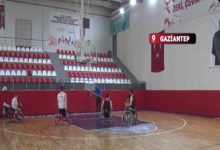 Görselde, bir spor salonunda tekerlekli sandalye basketbol takımı antrenman yapıyor. Oyuncular sahada dağılmış hâlde basket potasına doğru çalışırken, ortada bir antrenör onlara yönlendirme yapıyor. Tribünlerde kırmızı-beyaz koltuklar, yan duvarda Türk bayrağı ve yukarıda “GAZİANTEP” yazılı konum etiketi yer alıyor. Atmosfer, maç öncesi ciddi ve hazırlığa odaklı bir çalışma izlenimi veriyor.