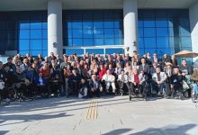 Büyük bir grubun, modern cam cepheli bir binanın önünde toplu fotoğraf verdiği görülüyor. Katılımcıların arasında tekerlekli sandalye kullanan bireyler de var. Herkes kameraya dönük, düzenli bir şekilde konumlanmış; ortam resmi bir buluşma ya da toplantı sonrası çekilen hatıra fotoğrafı havası taşıyor.