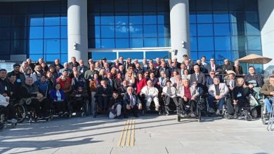 Büyük bir grubun, modern cam cepheli bir binanın önünde toplu fotoğraf verdiği görülüyor. Katılımcıların arasında tekerlekli sandalye kullanan bireyler de var. Herkes kameraya dönük, düzenli bir şekilde konumlanmış; ortam resmi bir buluşma ya da toplantı sonrası çekilen hatıra fotoğrafı havası taşıyor.