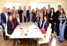 Bir toplantı odasında, farklı yaş gruplarından kalabalık bir ekip toplu fotoğraf çektiriyor. Masanın etrafında oturan ve ayakta duran katılımcılar, resmi ama samimi bir organizasyonun ardından objektife gülümseyerek poz veriyor.