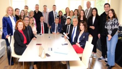 Bir toplantı odasında, farklı yaş gruplarından kalabalık bir ekip toplu fotoğraf çektiriyor. Masanın etrafında oturan ve ayakta duran katılımcılar, resmi ama samimi bir organizasyonun ardından objektife gülümseyerek poz veriyor.