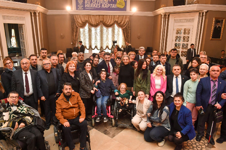 "Bu Şehirde Herkes Kaptan" yazılı bir pankartın önünde, tekerlekli sandalye kullanan bireylerin de dahil olduğu kalabalık bir grubun toplu fotoğrafıdır. Şık bir salon ortamında çekilen kare, kapsayıcı bir topluluk buluşmasını yansıtıyor.