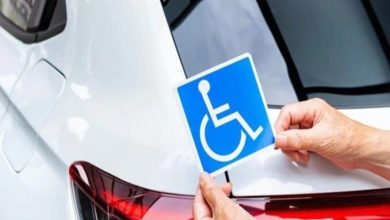 Bir elin, beyaz bir aracın arkasına mavi renkli engelli sembolü (tekerlekli sandalye ikonu) yapıştırdığı veya tuttuğu anı gösteren yakın çekim görsel. Fotoğraf, engelli bireylere yönelik araç kullanım haklarını veya muafiyetlerini temsil eden sembolik bir anlatıma sahip.