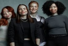 Yan yana duran dört genç kadın, kameraya gülümseyerek poz veriyor. Farklı saç stilleri ve kıyafetlerle, sade arka plan önünde samimi bir grup portresi oluşturuyorlar.
