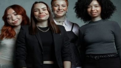 Yan yana duran dört genç kadın, kameraya gülümseyerek poz veriyor. Farklı saç stilleri ve kıyafetlerle, sade arka plan önünde samimi bir grup portresi oluşturuyorlar.