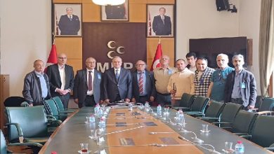 Toplantı salonunda çekilen fotoğrafta, bir grup ziyaretçi ve yetkili uzun bir toplantı masasının önünde yan yana poz veriyor. Arka planda MHP logosu, Türk bayrakları ve siyasi liderlerin portreleri yer alıyor. Görüşme resmi bir atmosferde gerçekleşiyor.