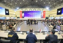 Geniş ve modern bir konferans salonunda, AFAD logosunun bulunduğu büyük ekranların önünde çok sayıda yetkili ve katılımcı, afet farkındalığına yönelik bir toplantı için yarım daire şeklinde oturma düzeninde bir araya gelmiş.