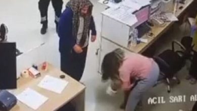 Bir güvenlik kamerası görüntüsünde, hastane benzeri bir ortamda küçük bir çocuğa müdahale eden bir kadın görülüyor. Kadın eğilmiş şekilde çocuğa yardım etmeye çalışırken, çevrede bulunan diğer kişiler durumu endişeyle izliyor. Görüntü, acil bir sağlık müdahalesi anını yansıtıyor.