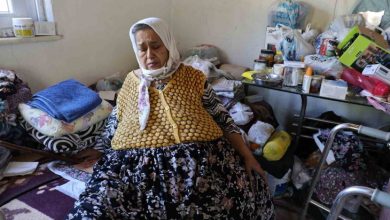 Mütevazı bir ev ortamında yaşlı bir kadın yerde otururken görülüyor. Etrafında günlük yaşam eşyaları, ilaçlar ve yürüme desteği sağlayan ekipmanlar bulunuyor. Görsel, sağlık sorunları nedeniyle zor koşullarda sürdürülen yaşam mücadelesini yansıtıyor.