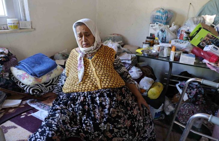 Mütevazı bir ev ortamında yaşlı bir kadın yerde otururken görülüyor. Etrafında günlük yaşam eşyaları, ilaçlar ve yürüme desteği sağlayan ekipmanlar bulunuyor. Görsel, sağlık sorunları nedeniyle zor koşullarda sürdürülen yaşam mücadelesini yansıtıyor.