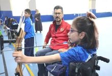 Kapalı bir spor salonunda, tekerlekli sandalyede bulunan bir kız çocuğu ok atışı yaparken görülüyor. Yanında, ona rehberlik eden kırmızı montlu bir antrenör dikkatle süreci izliyor. Arka planda başka çocuklar da okçuluk çalışması yapıyor. Görsel, engelli çocukların sporla buluştuğu, destekleyici ve motive edici bir anı yansıtıyor.