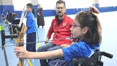 Kapalı bir spor salonunda, tekerlekli sandalyede bulunan bir kız çocuğu ok atışı yaparken görülüyor. Yanında, ona rehberlik eden kırmızı montlu bir antrenör dikkatle süreci izliyor. Arka planda başka çocuklar da okçuluk çalışması yapıyor. Görsel, engelli çocukların sporla buluştuğu, destekleyici ve motive edici bir anı yansıtıyor.