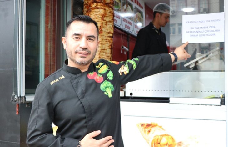 Önünde “İkramda Engel Yok” yazan afişi gösteren bir aşçı ve arka planda döner tezgâhı.