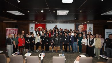 Görselde, bir konferans salonunda büyük bir grup katılımcı sahne önünde toplu fotoğraf çekiliyor. Arkada Türk bayrağı ve Atatürk portresi yer alıyor. Katılımcılar çoğunlukla yetişkinlerden oluşuyor; resmi ve yarı resmi kıyafetlerle poz veriyorlar. Salonun ön sırasındaki koltuklar boş, önde küçük bir çocuk kameraya doğru bakıyor. Genel atmosfer resmî, akademik ve toplu etkinlik havasında.