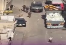 Yüksekten çekilmiş bir sokak görüntüsünde, araçların bulunduğu dar bir yolda birkaç kişi koşuşturuyor. Bir kamyon ve otomobiller yol üzerinde dururken, çevrede dağınık eşyalar dikkat çekiyor. Sahne, ani gelişen bir olay ya da gerginlik anını andırıyor.