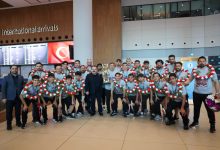 Uluslararası geliş terminalinde çekilen bu fotoğrafta, boyunlarında kırmızı-beyaz çiçeklerden oluşan çelenkler taşıyan genç sporcular ve antrenörleri, kazandıkları kupayla birlikte gururla poz verirken görülüyor.