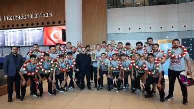 Uluslararası geliş terminalinde çekilen bu fotoğrafta, boyunlarında kırmızı-beyaz çiçeklerden oluşan çelenkler taşıyan genç sporcular ve antrenörleri, kazandıkları kupayla birlikte gururla poz verirken görülüyor.