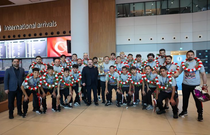 Uluslararası geliş terminalinde çekilen bu fotoğrafta, boyunlarında kırmızı-beyaz çiçeklerden oluşan çelenkler taşıyan genç sporcular ve antrenörleri, kazandıkları kupayla birlikte gururla poz verirken görülüyor.