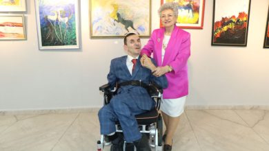 Şık Vahit Sonay Yazgan tekerlekli sandalyede otururken, yanında duran pembe ceketli emekli sınıf öğretmeni Bedriye Yazgan, oğlunun omzuna sevgiyle dokunarak birlikte sanat galerisi ortamında poz veriyor.