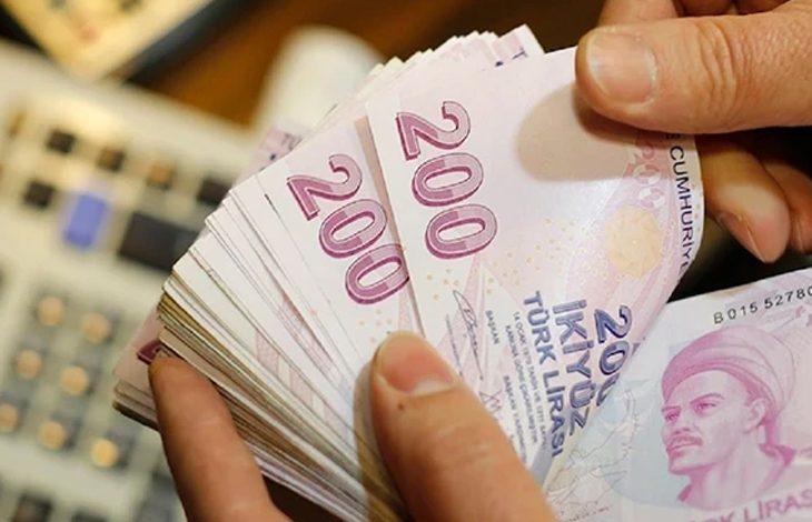 Bir kişinin elinde deste hâlinde 200 TL banknotlar var; arka planda bulanık bir hesap makinesi görülüyor.