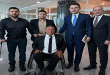 Resmî bir bina içinde çekilen fotoğrafta takım elbiseli beş kişi birlikte poz veriyor. Ortada tekerlekli sandalyede oturan bir kişinin yanında duran grup, resmi bir görüşme ya da ziyaret sonrası objektife bakıyor.