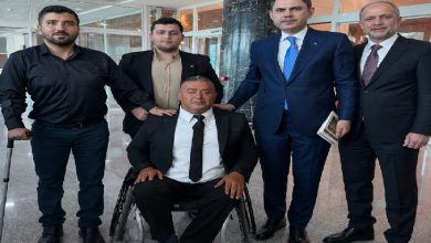 Resmî bir bina içinde çekilen fotoğrafta takım elbiseli beş kişi birlikte poz veriyor. Ortada tekerlekli sandalyede oturan bir kişinin yanında duran grup, resmi bir görüşme ya da ziyaret sonrası objektife bakıyor.