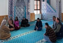 Bir caminin iç mekânında, turkuaz desenli halı üzerinde kadın ve erkeklerden oluşan bir grup daire şeklinde oturmuş. Duvarlar mavi-beyaz çinilerle süslü, sağda minber ve pencere görünüyor. Ortam sakin, sohbet ya da toplantı havası var.